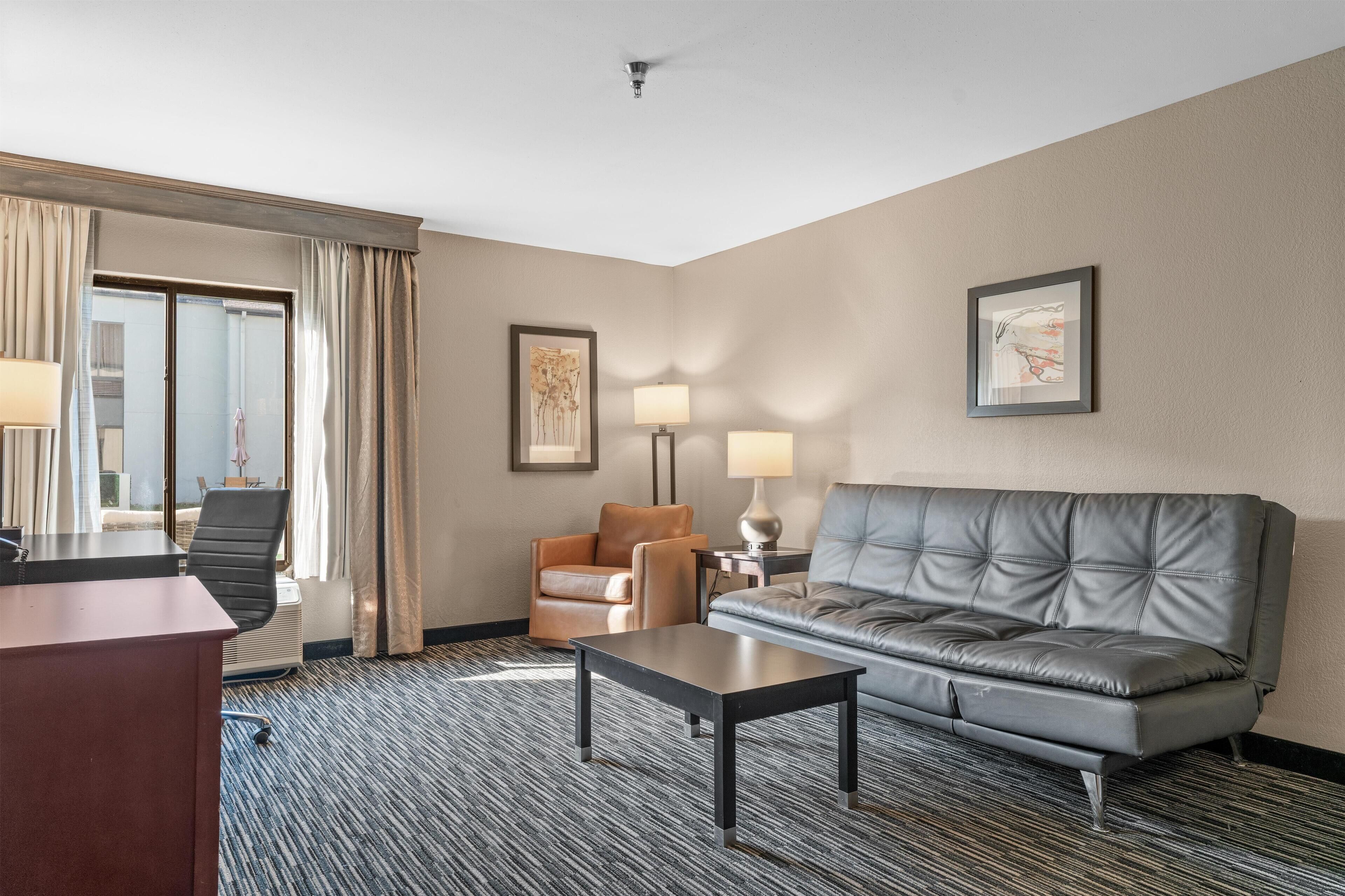 A hotel room with a leather sofa, a small table, a chair, a desk, and a large window with curtains.