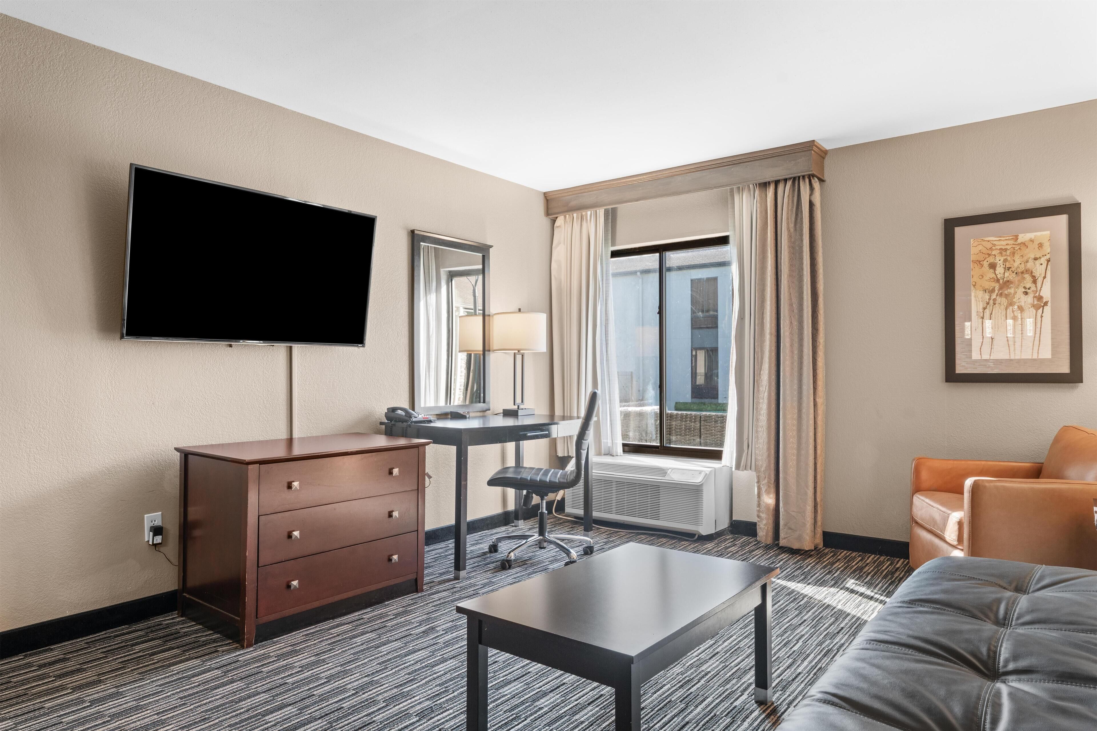 A hotel room with a flat-screen TV, a desk with a chair, a sofa, a dresser, and a large window with curtains.