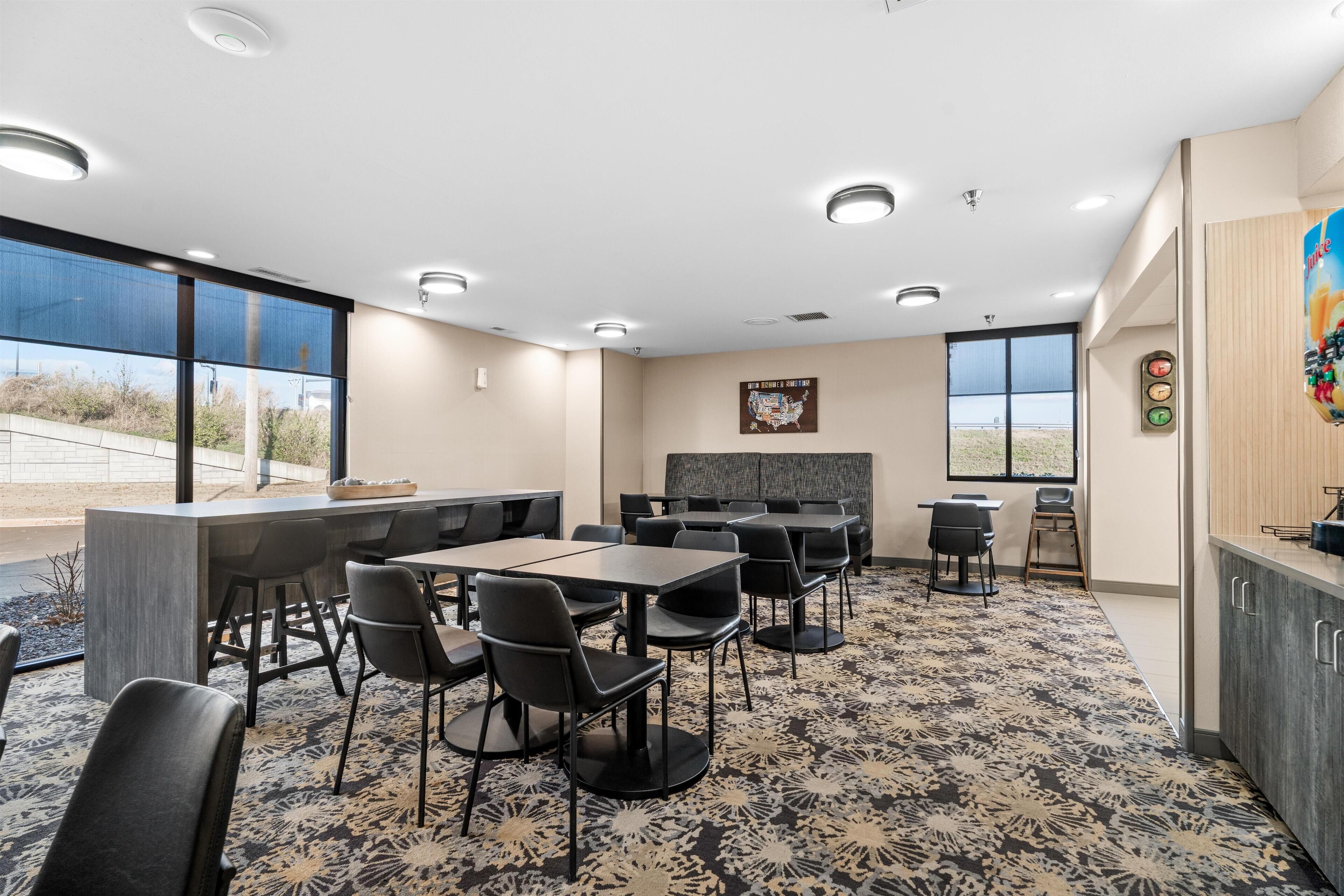 A conference room with a large table and chairs, a kitchen area, and large windows offering a view of the outdoors.