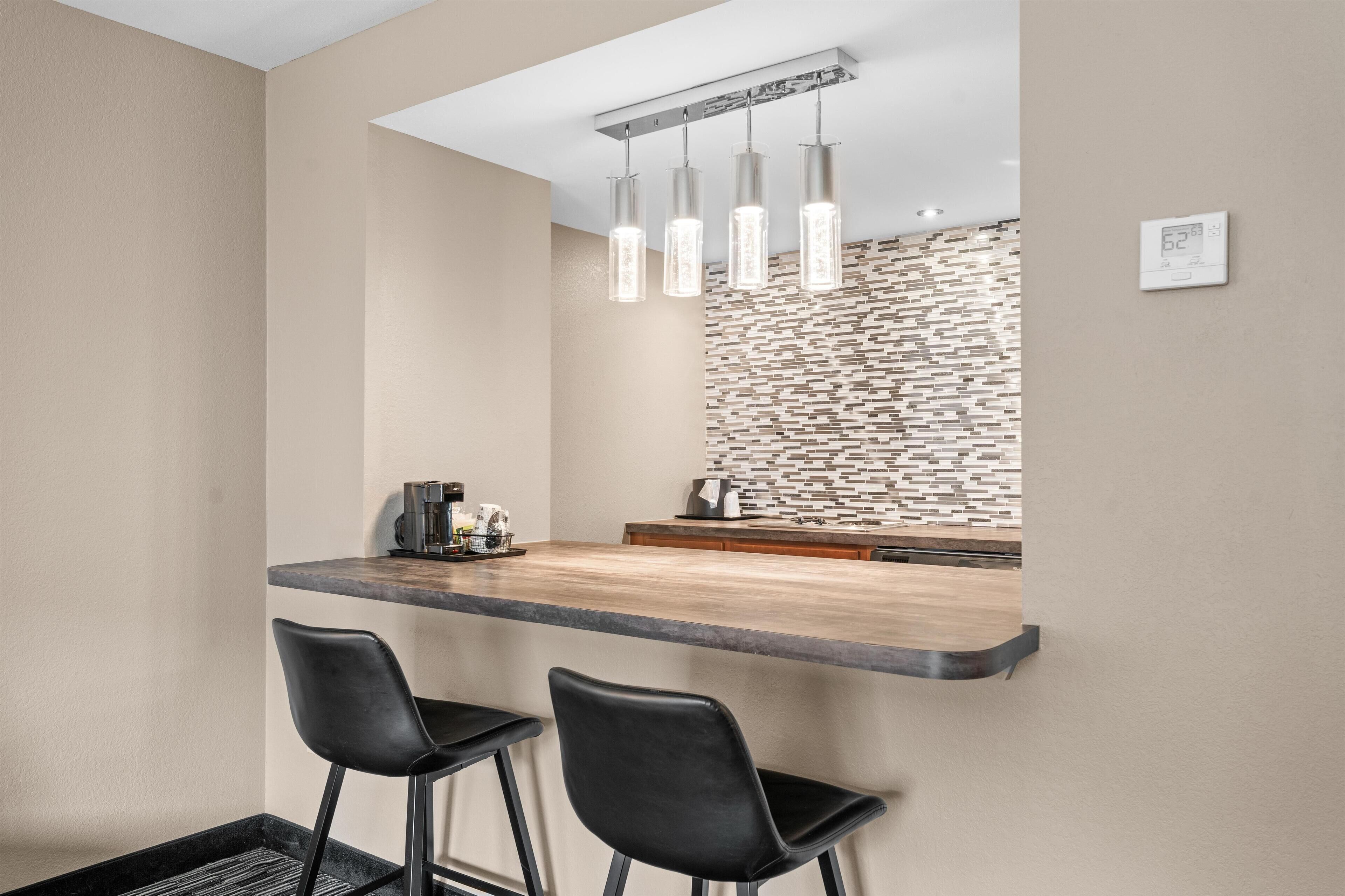 Una cocina moderna con encimera de madera, dos taburetes negros, iluminación colgante y un salpicadero con diseño.