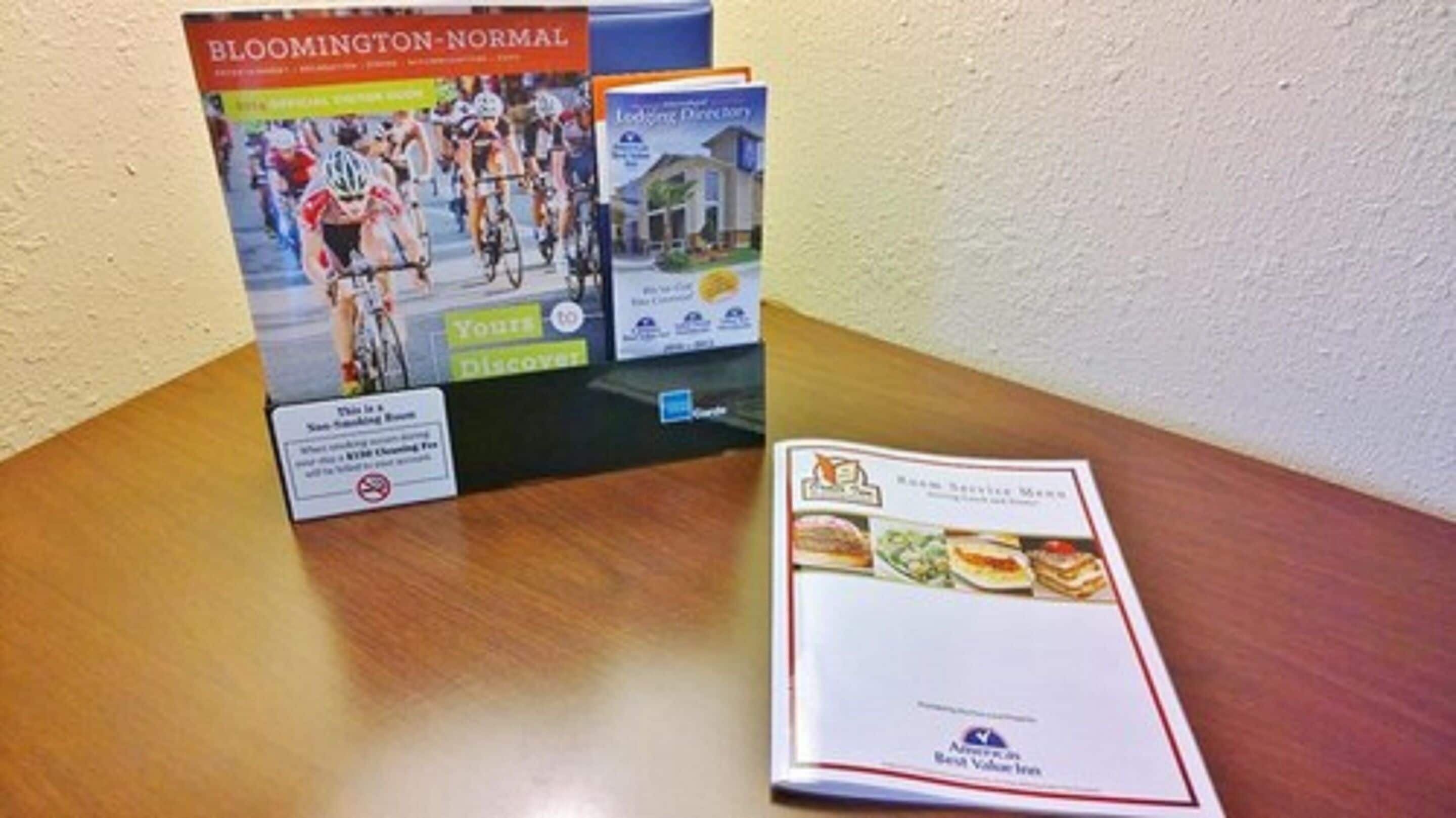 Un folleto con el título 'Bloomington-Normal' que muestra un evento de ciclismo, un libro 'Guía de Alojamientos' y un folleto 'Menú de Servicio a la Habitación' sobre una mesa.