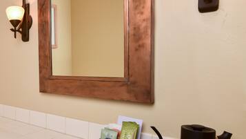 Une salle de bain avec un grand miroir, deux lampes fixées au mur, un plateau avec des tasses à café, une bouilloire et un paquet de capsules de café.