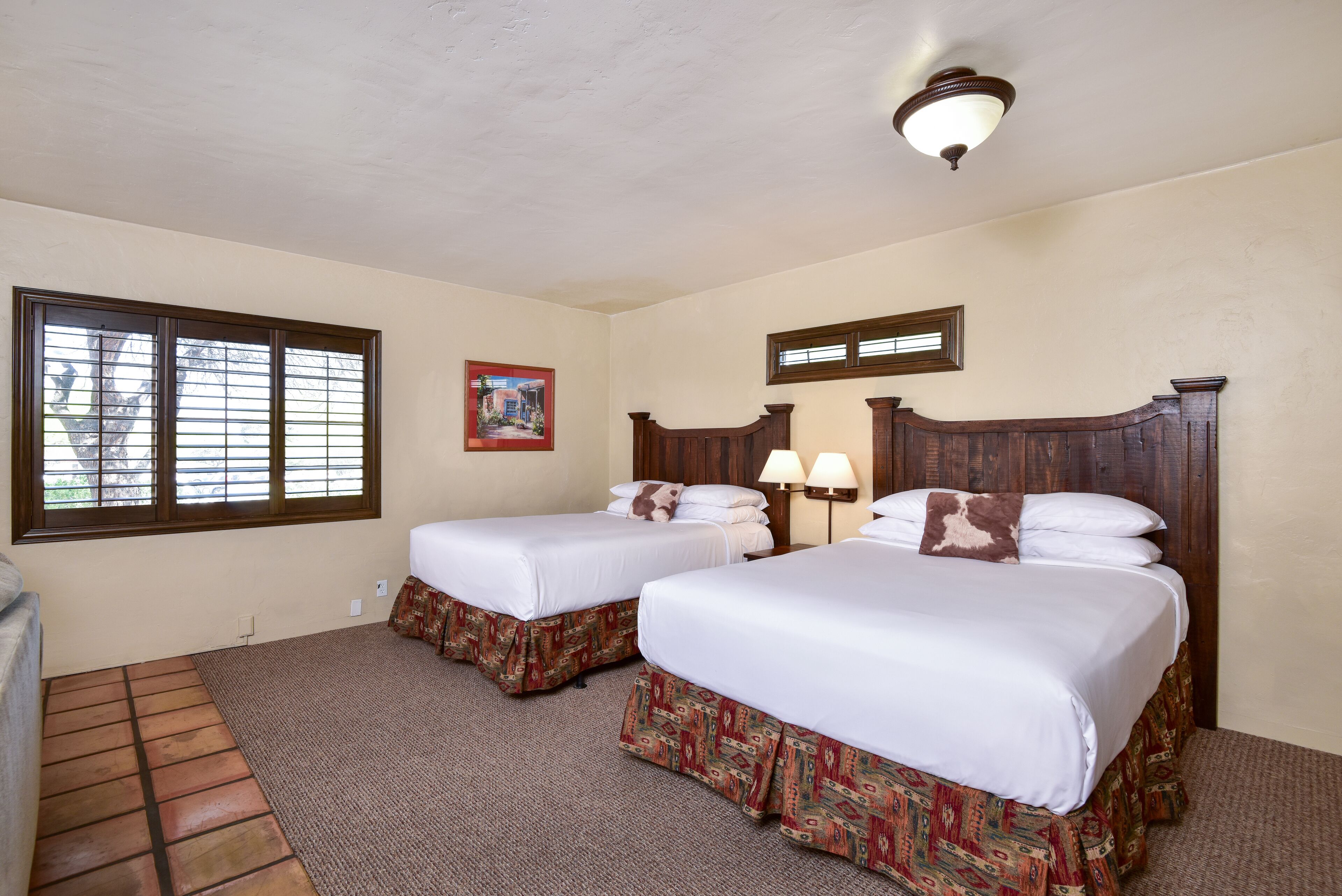 A hotel room with two beds, a large window, a painting, and a ceiling light fixture.