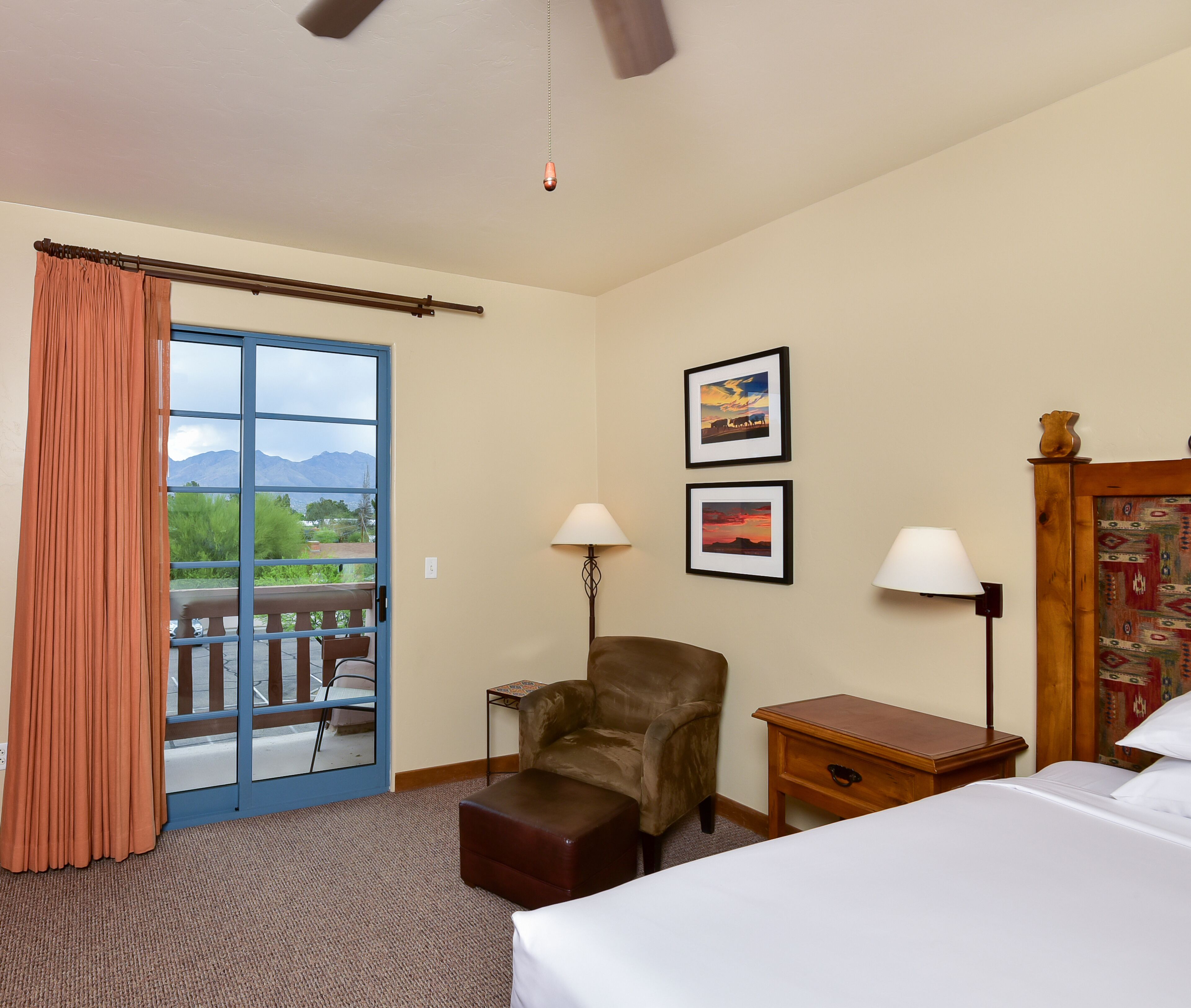 A hotel room with a bed, a chair, a small table, two wall-mounted lamps, a wooden nightstand, and a view of mountains through a glass door.