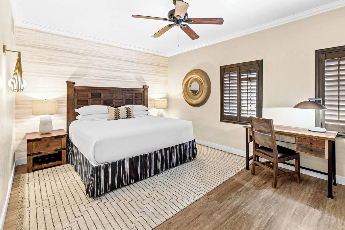 Une chambre à coucher comprenant un lit, un bureau, une chaise, un ventilateur de plafond et deux fenêtres équipées de stores.