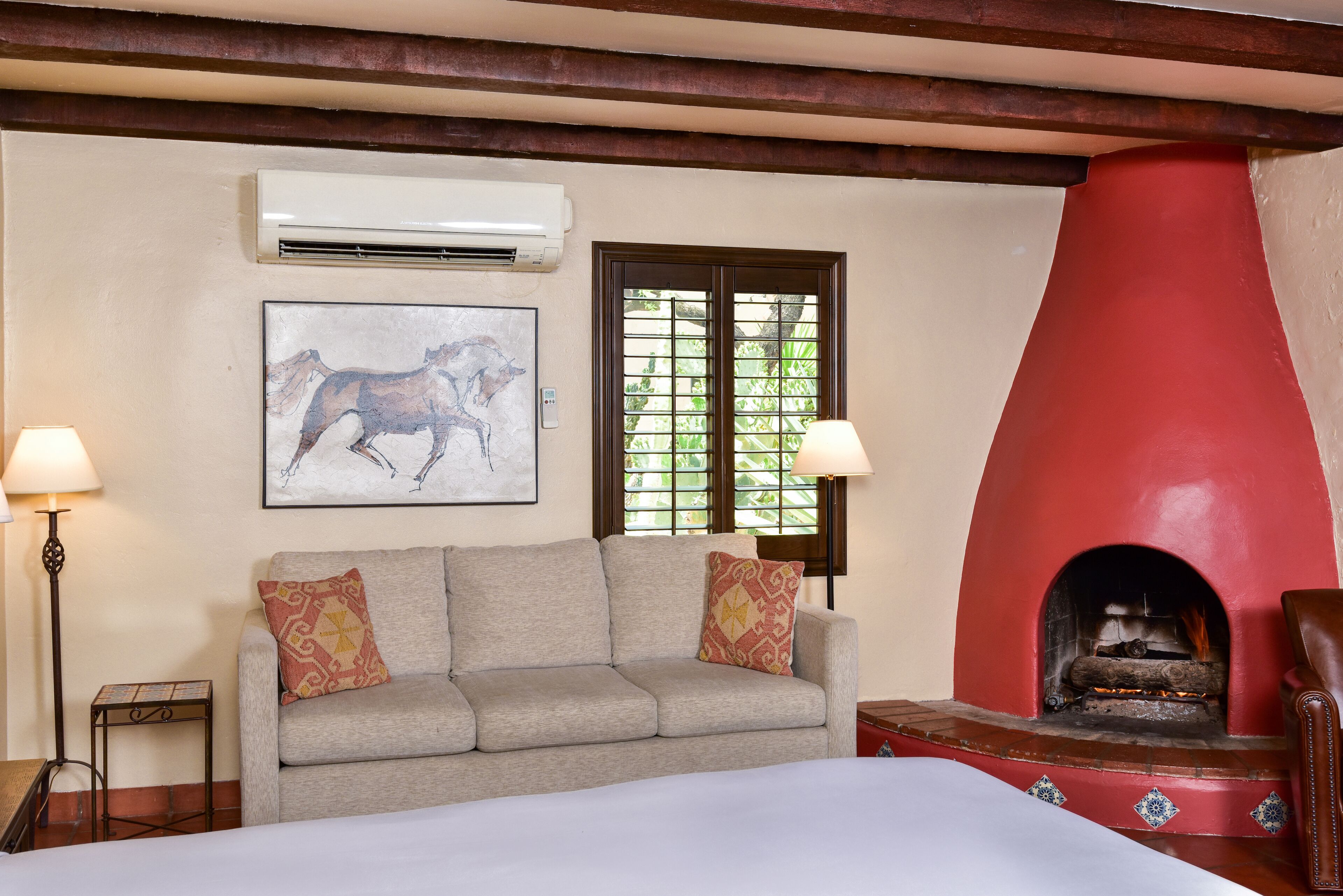 A room with a beige sofa, two pillows, a red fireplace, an air conditioning unit, and a framed picture on the wall.