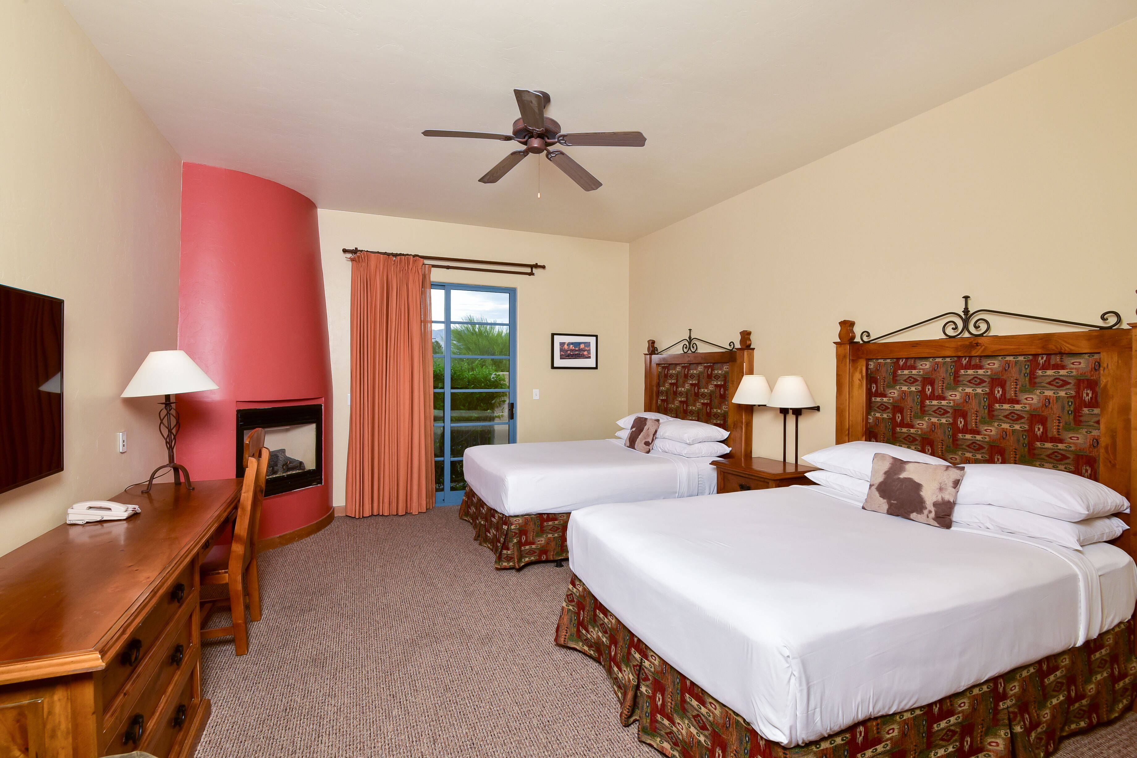 A hotel room with two beds, a desk, a chair, a ceiling fan, a fireplace, and a window with curtains.