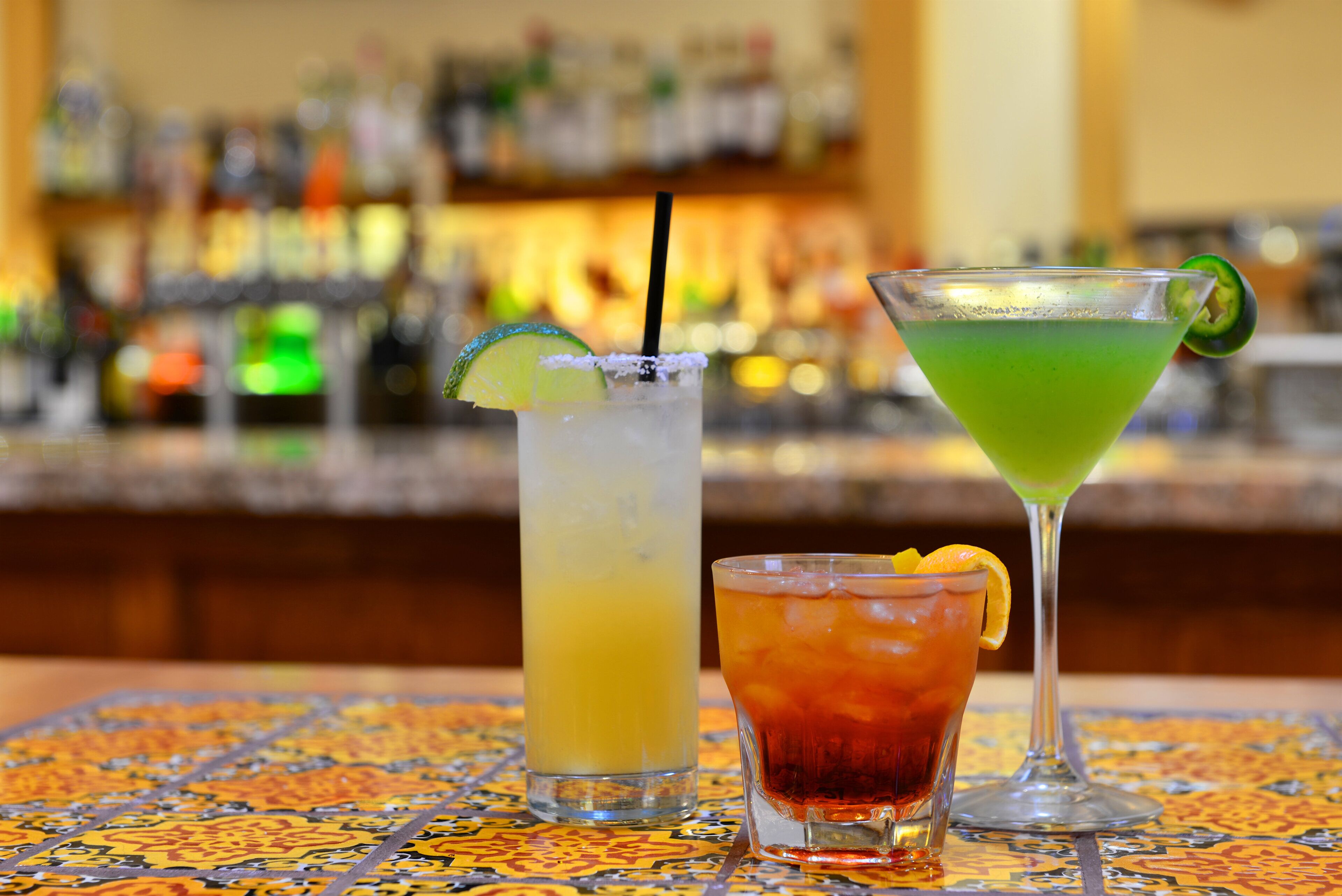 Three cocktails on a bar counter with a blurred background of bottles.