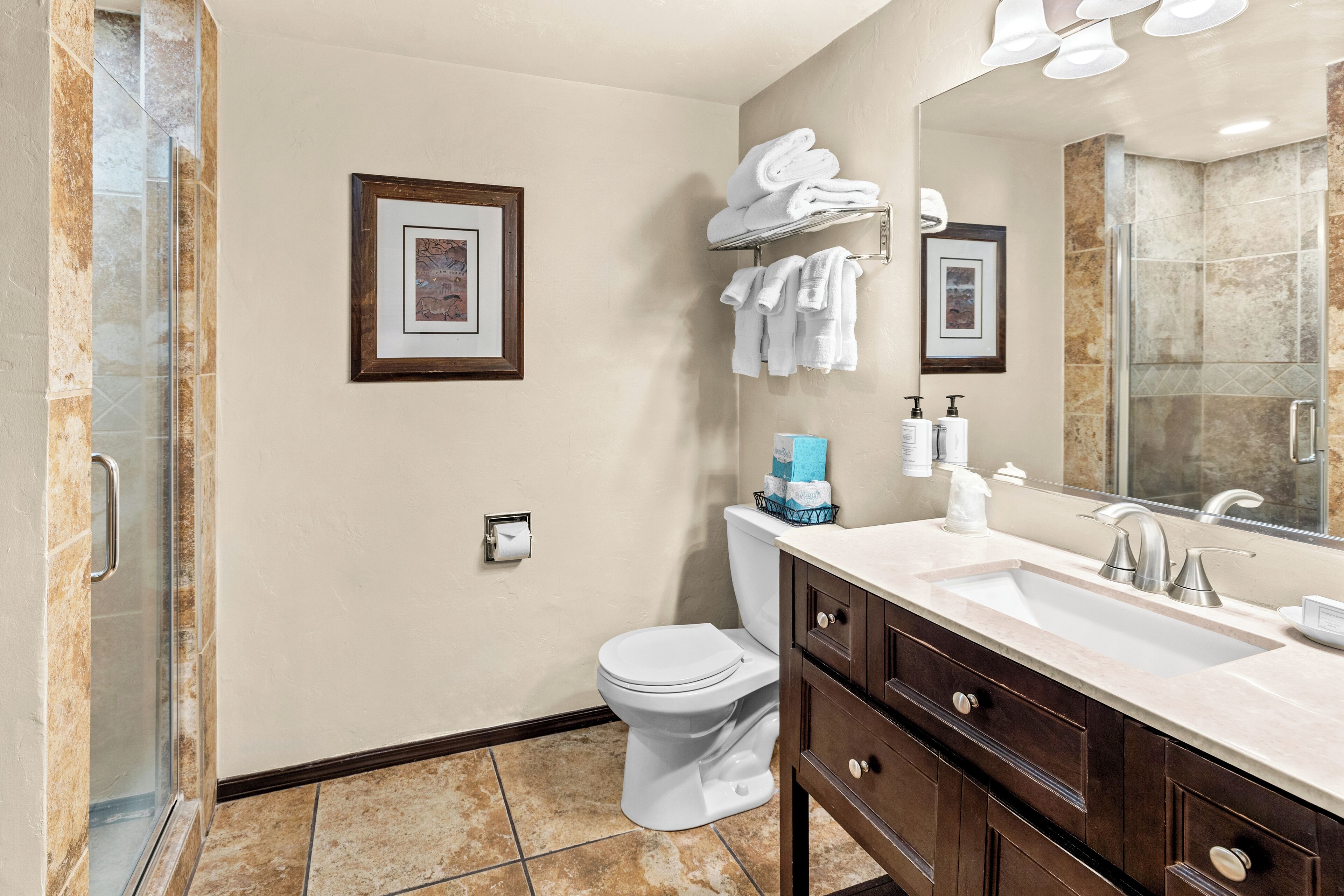 A modern bathroom with a glass shower enclosure, a white toilet, a vanity with a sink and mirror, and two framed pictures on the wall.