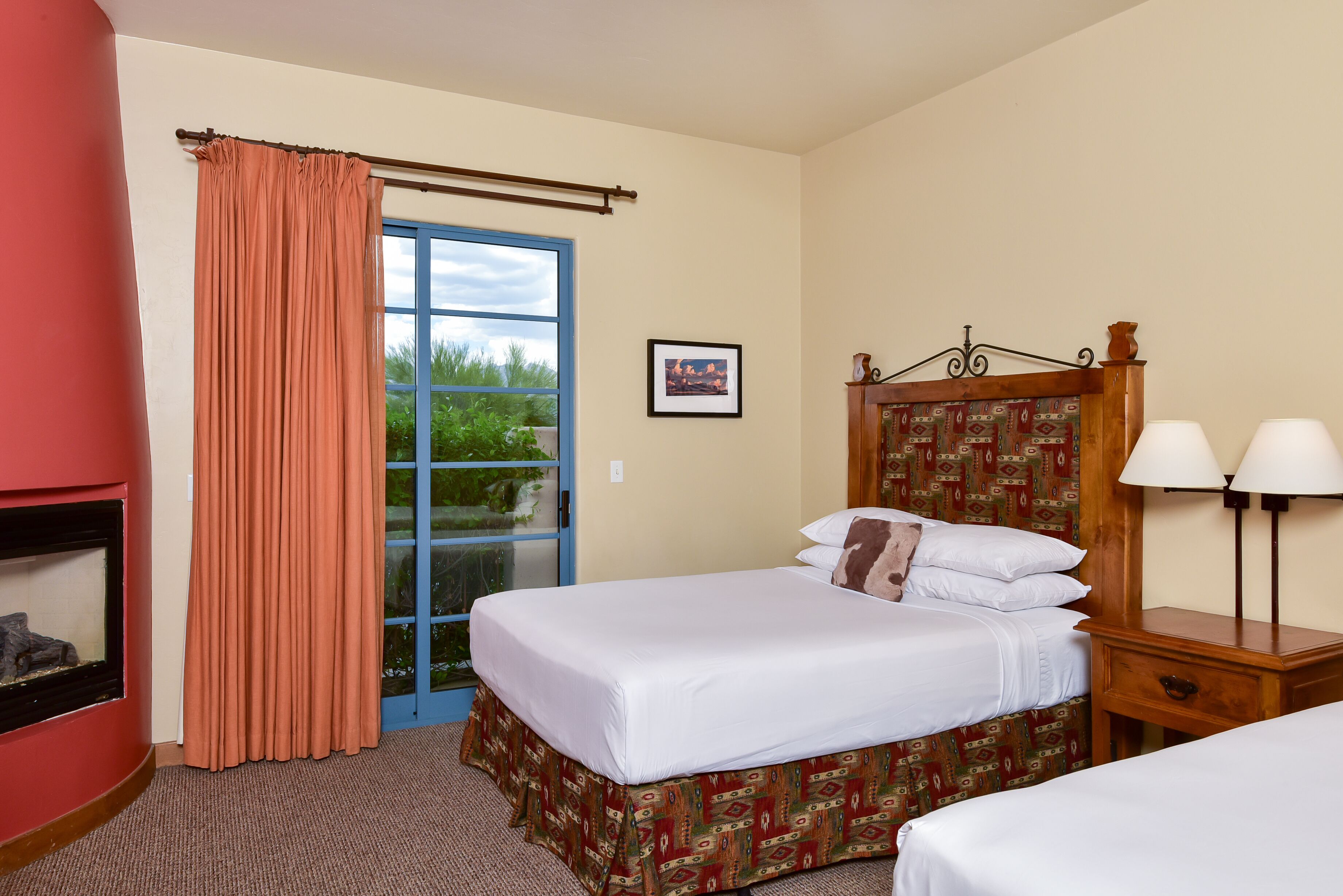 A hotel room with a bed, a nightstand with lamps, a window with curtains, and a fireplace.