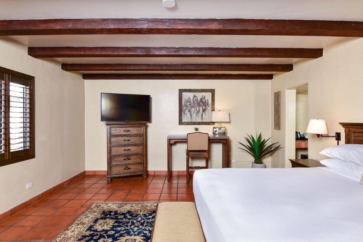 A bedroom with a bed, desk, chair, television, and a potted plant.