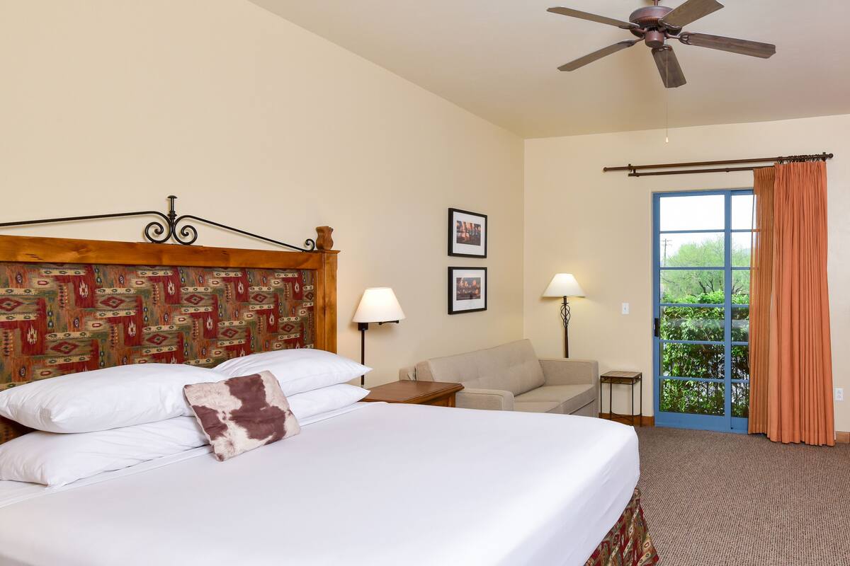 Une chambre d’hôtel avec un lit, un ventilateur de plafond, un canapé, une petite table, une lampe et une vue sur la verdure par la fenêtre.