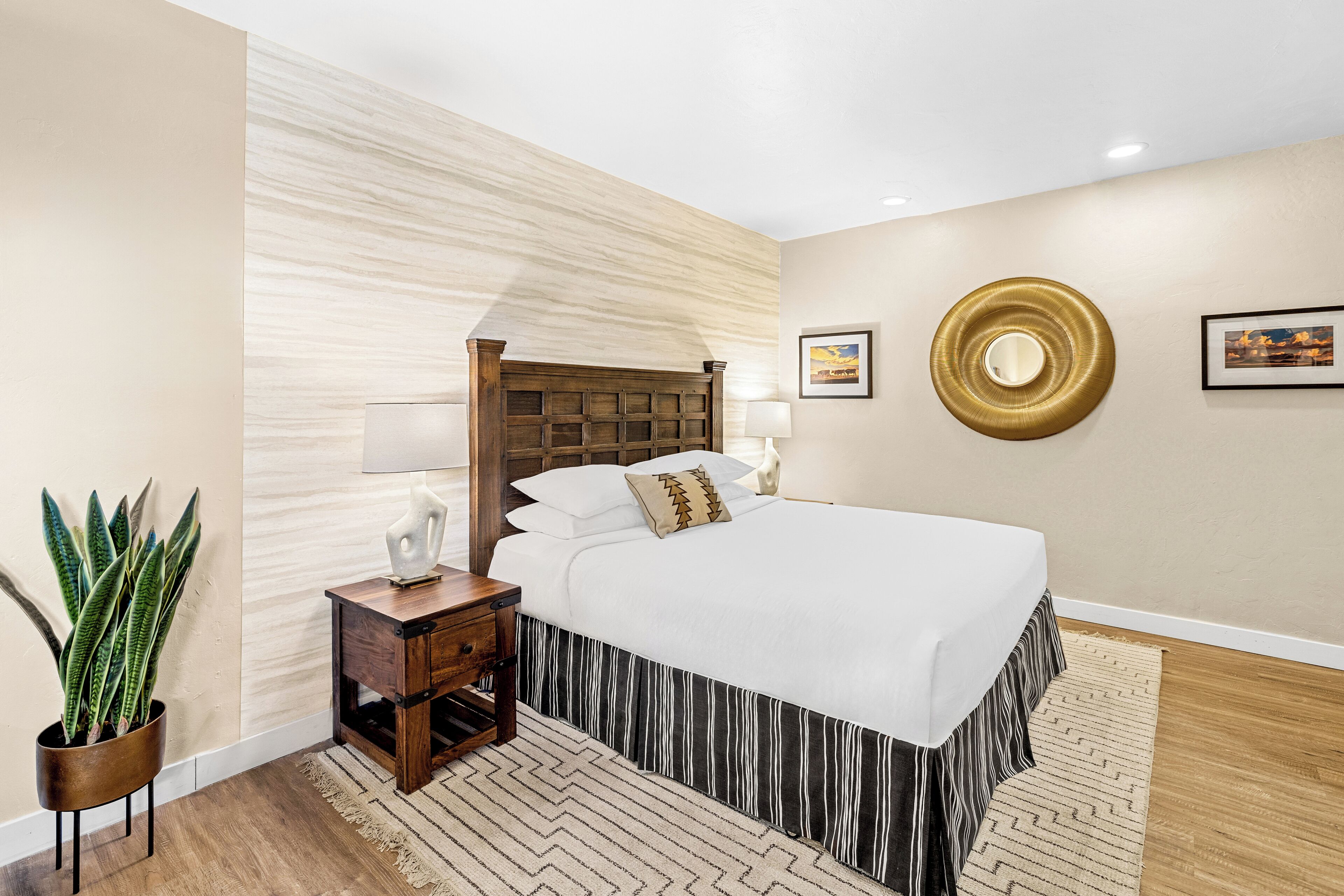 Quarto com cabeceira de madeira, cama com roupa de cama branca, mesa de cabeceira com abajur, planta em vaso e tapete estampado.