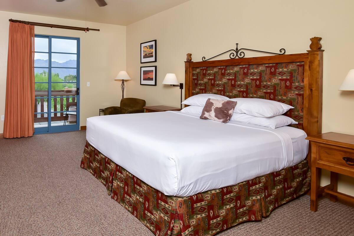 Une chambre d’hôtel avec un grand lit, un ventilateur de plafond, un bureau avec une lampe, une chaise et une vue sur les montagnes, visible par une porte coulissante.
