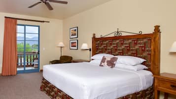 A hotel room with a large bed, a ceiling fan, a desk with a lamp, a chair, and a view of mountains through a sliding door.