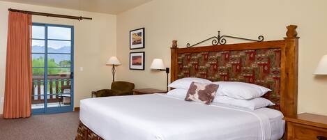 Habitación de hotel con una cama grande, un ventilador de techo, un escritorio con lámpara, una silla y vistas a montañas a través de una puerta corredera.