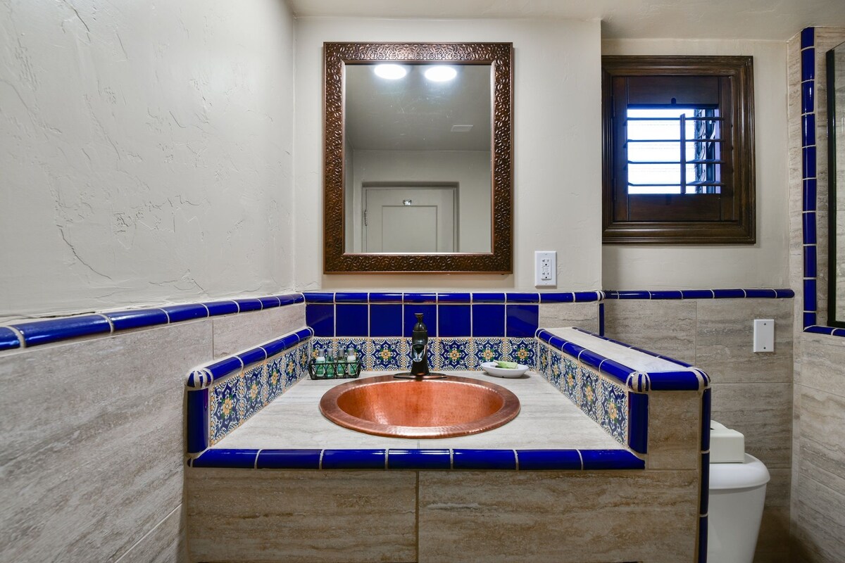 A bathroom with a copper sink, blue-tiled walls, a mirror, and a window with bars.