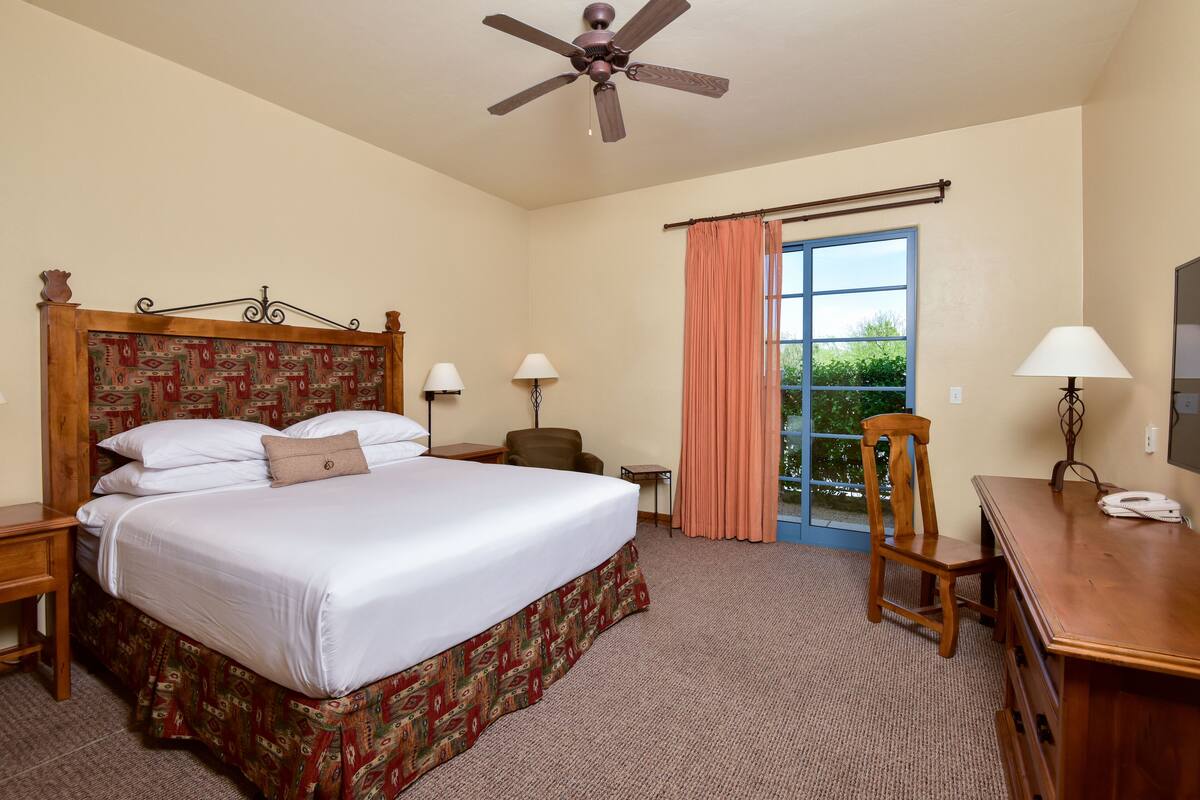 Une chambre d’hôtel avec un grand lit, un bureau, une chaise, un ventilateur de plafond et une fenêtre avec des rideaux.