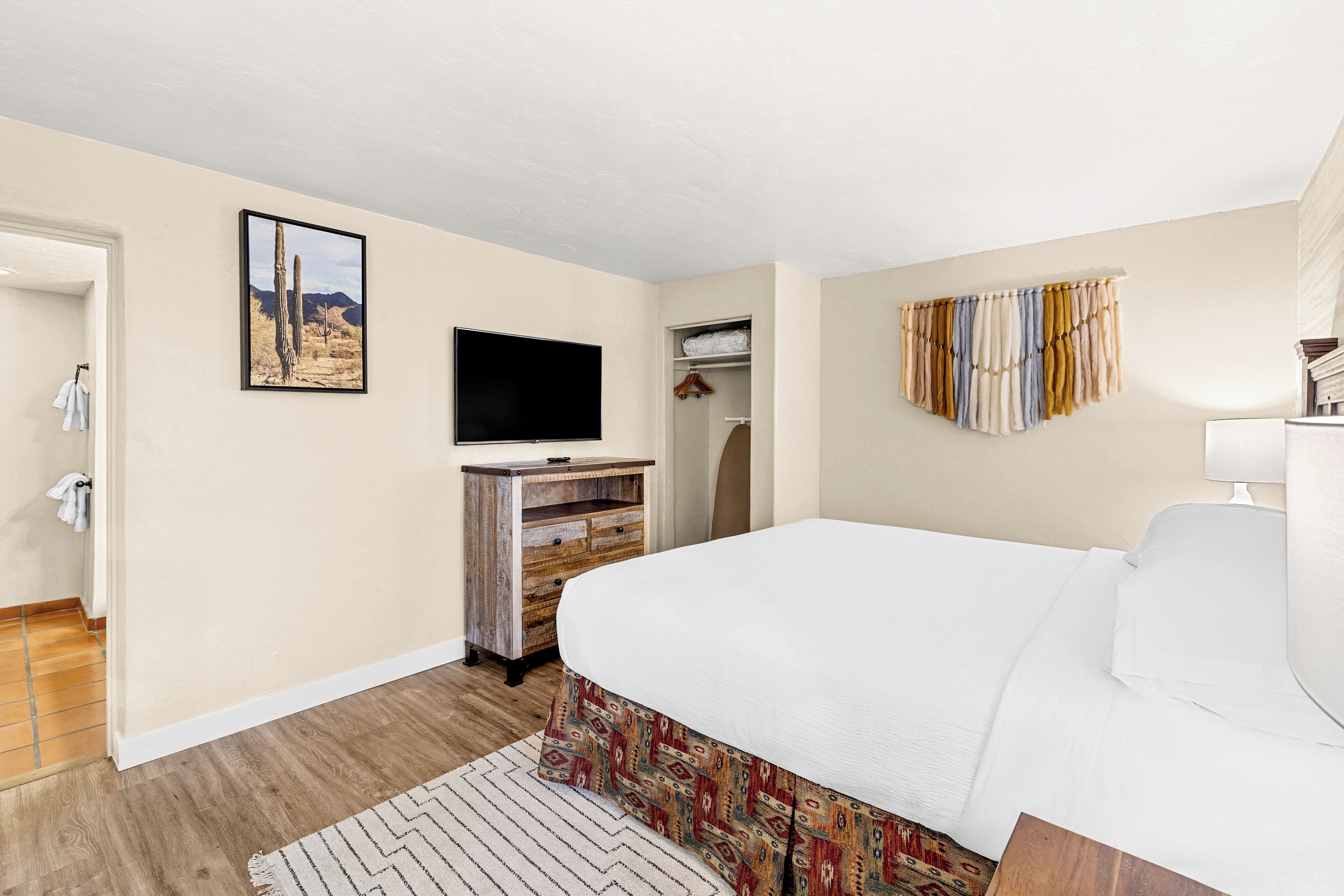 A hotel room with a bed, a TV, a wooden dresser, a patterned rug, and a desert-themed wall art.
