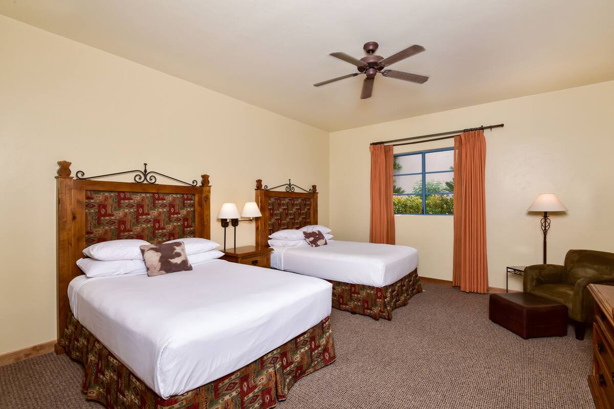 Une chambre d’hôtel avec deux lits simples, un ventilateur de plafond, une fenêtre avec des rideaux, une lampe et une chaise.