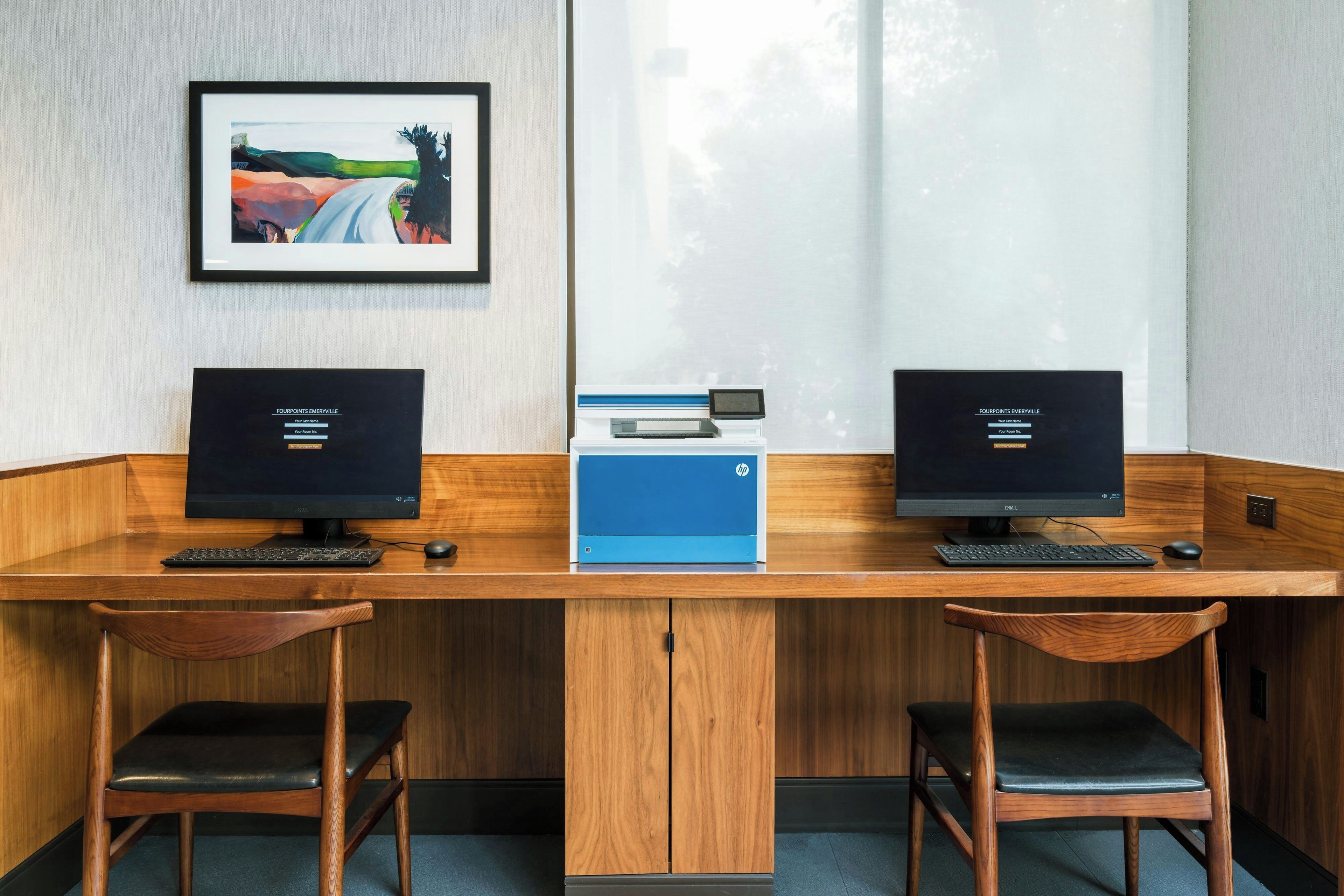 Un espacio de trabajo con dos escritorios, cada uno con un monitor de computadora, teclado y ratón. Hay dos sillas, un cuadro enmarcado en la pared y una impresora azul en el centro.
