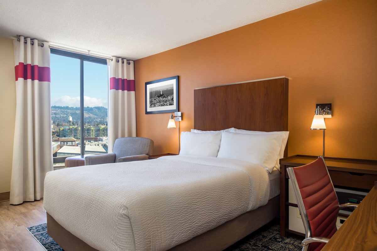 Ein Hotelzimmer mit einem großen Bett, einem Schreibtisch, einem Sessel und Blick auf die Stadt, sowie einem gerahmten Bild an der Wand.