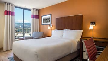 A hotel room with a large bed, a desk, a chair, a view of the city, and a framed picture on the wall.
