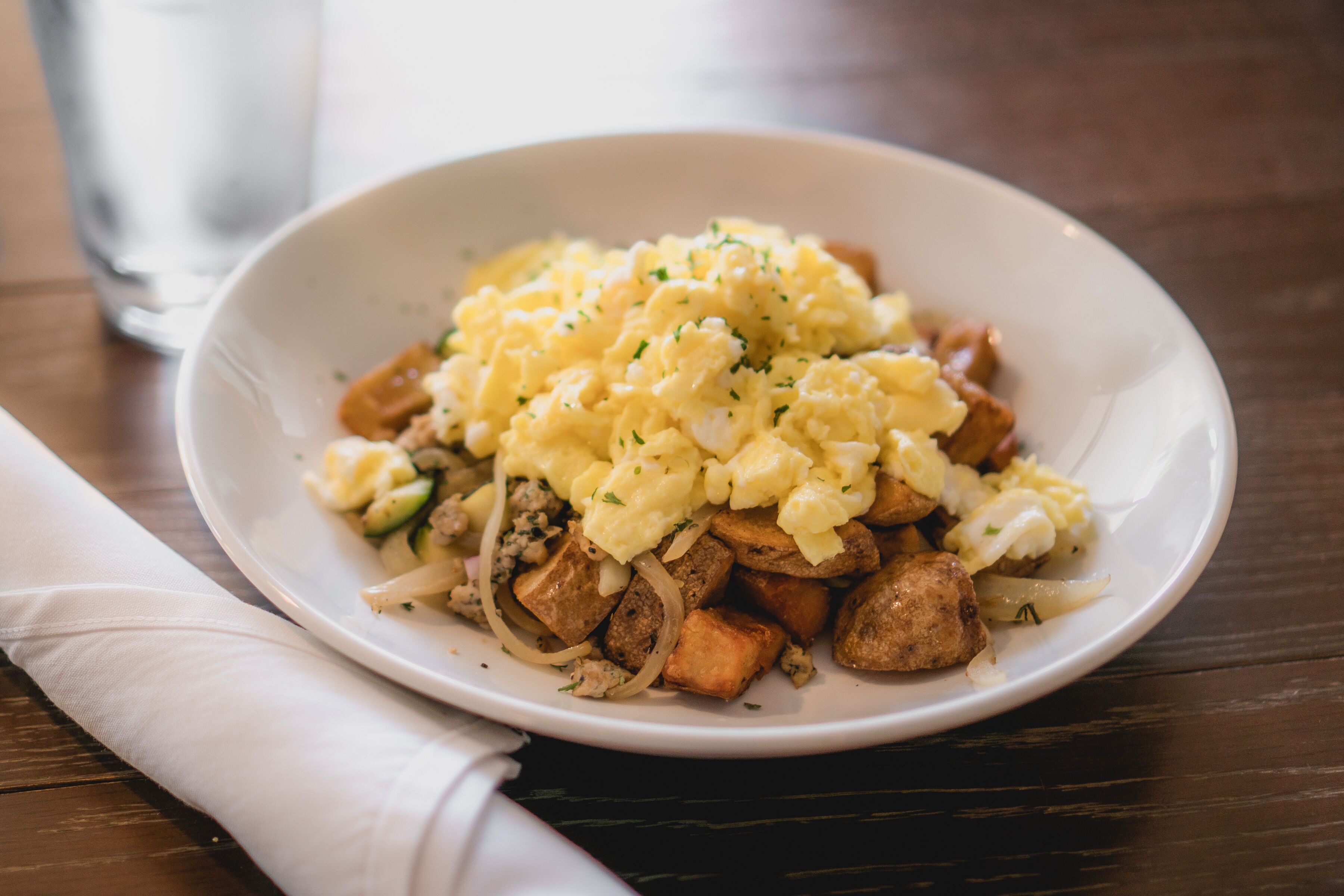 A white plate with scrambled eggs, diced potatoes, and onions on a wooden table.