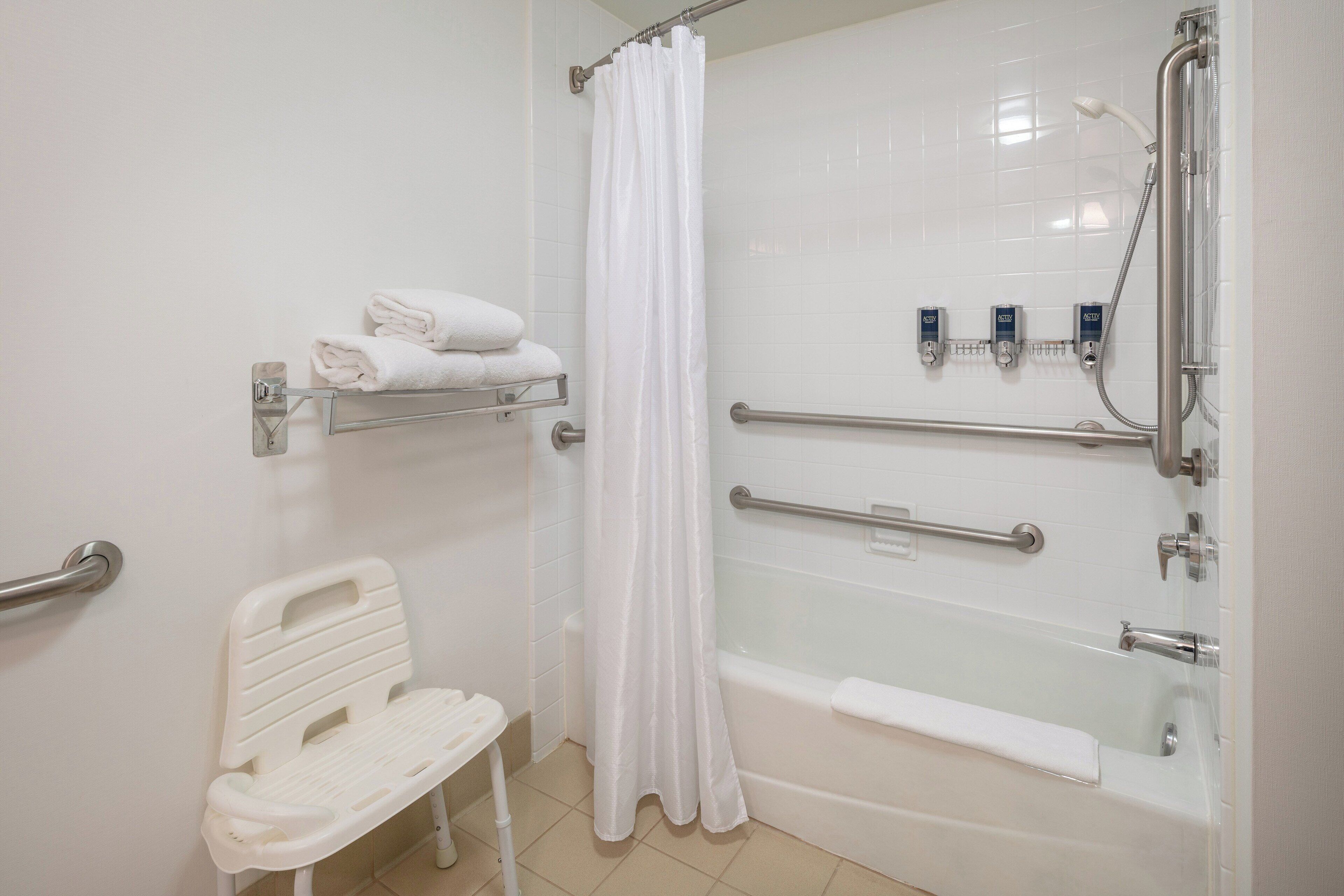 A bathroom with a white bathtub, a shower with a glass door, a white chair, and a towel rack with folded towels.