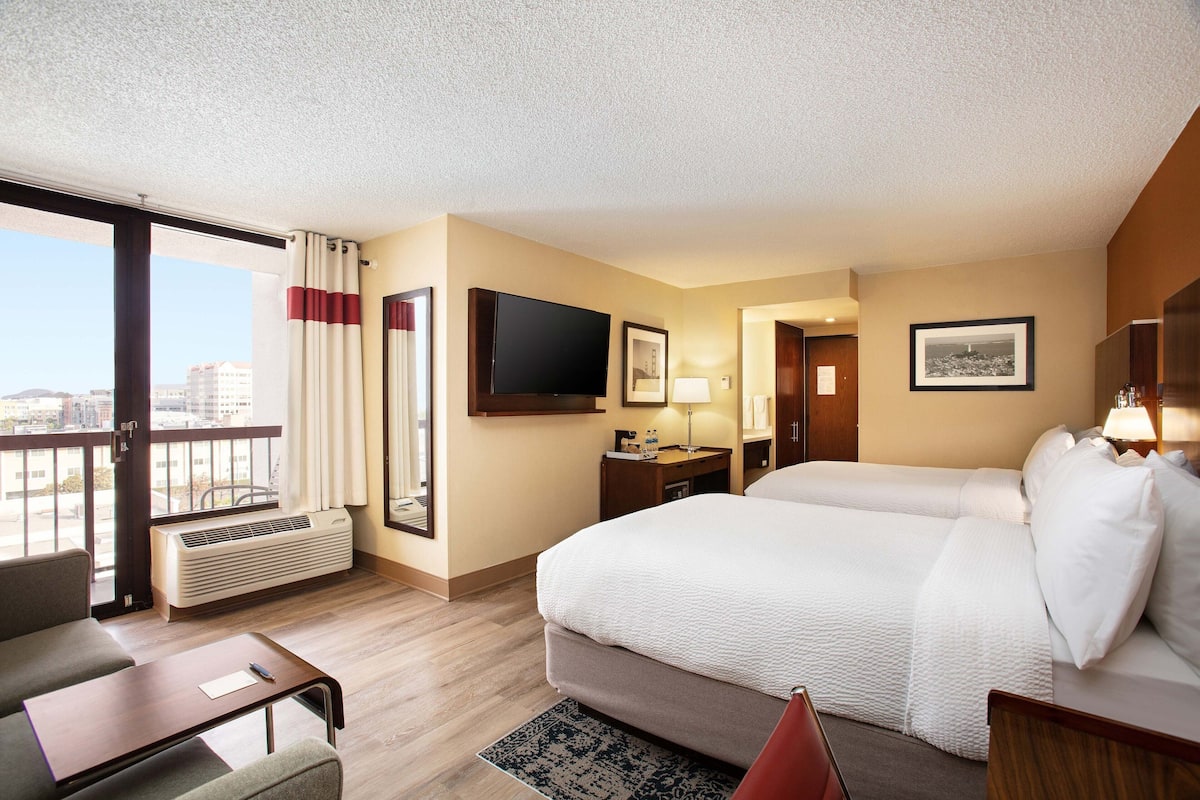 Ein Hotelzimmer mit einem großen Bett, einem Schreibtisch mit Stuhl, einem Fernseher und einem Fenster mit Vorhängen.