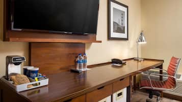 A hotel room with a wooden desk, a flat-screen TV, a coffee machine, bottled water, a chair, and a framed picture on the wall.