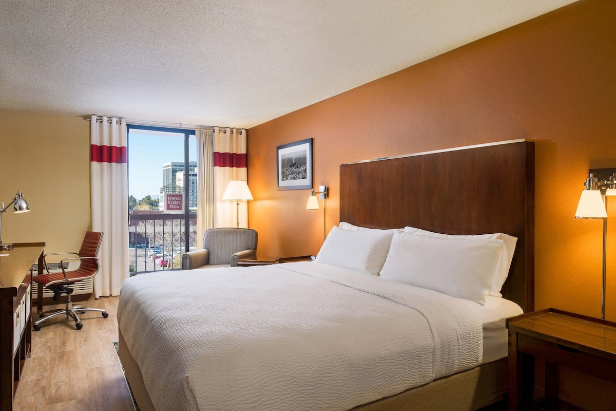 Ein Hotelzimmer mit einem großen Bett, einem Schreibtisch mit Stuhl, einer Lampe und Blick auf die Stadt durch das Fenster.