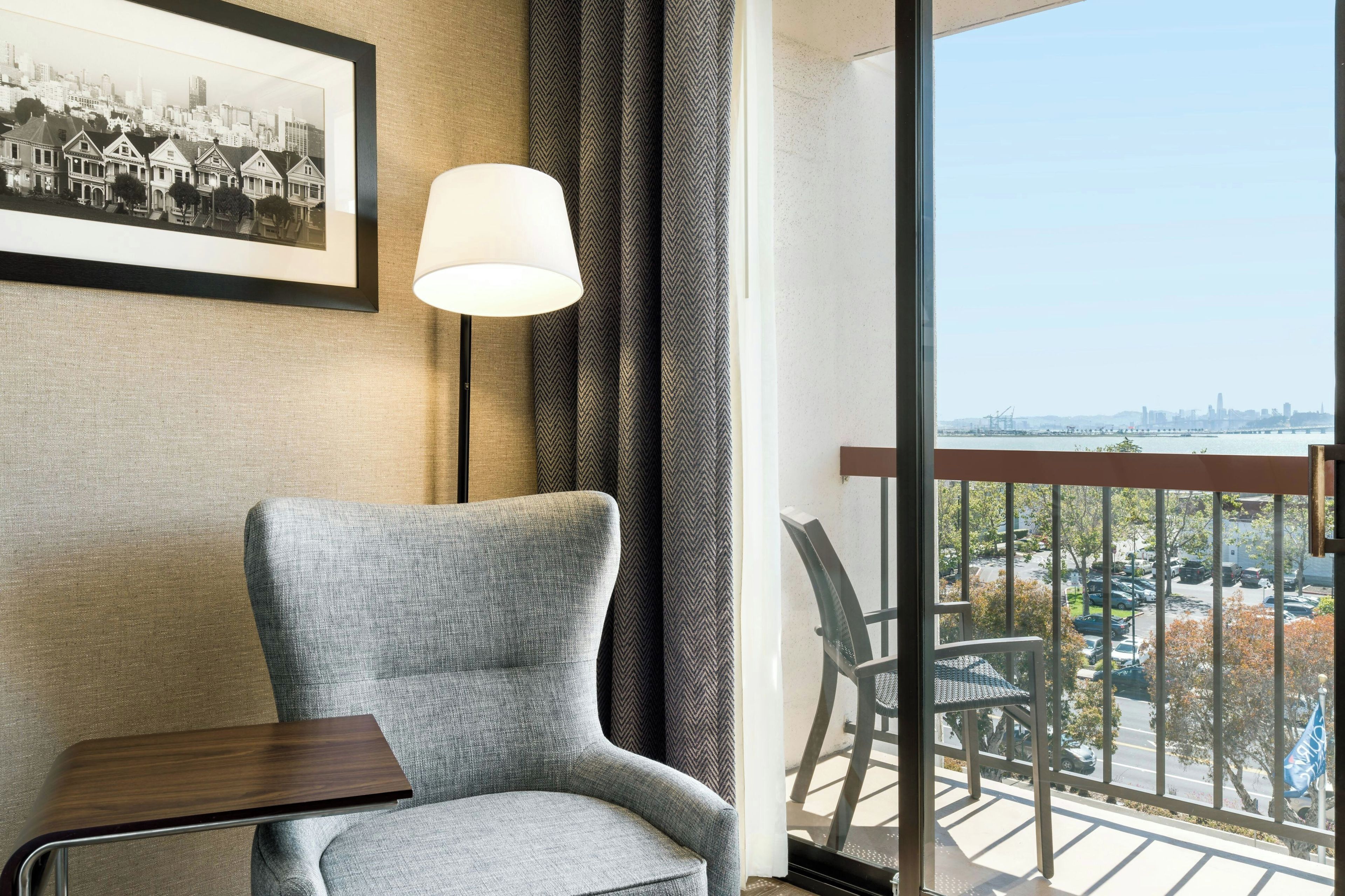 A room with a grey armchair, a small wooden table, a framed black and white photo, a patterned curtain, and a balcony with a view of a cityscape.