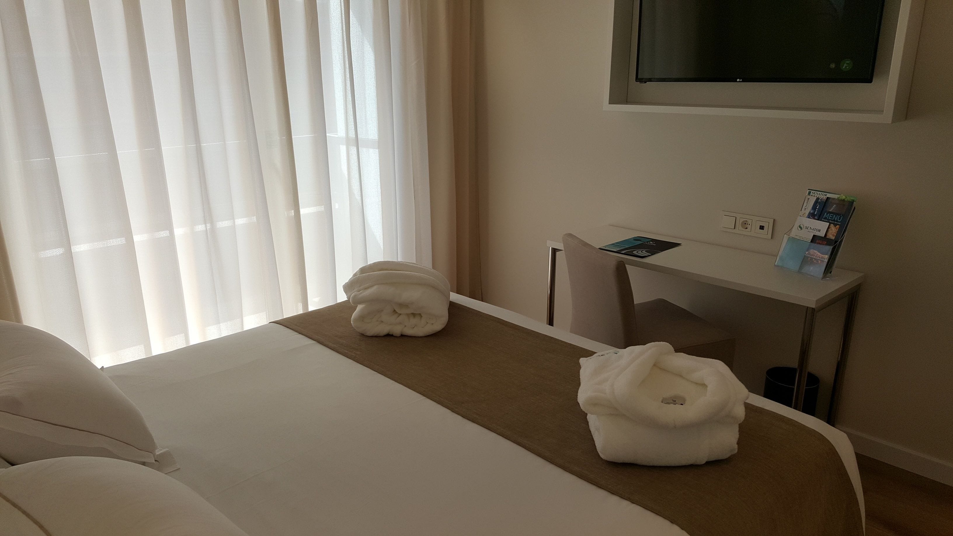Una habitación de hotel con cama, escritorio, televisor y ventana con cortinas.
