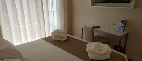 Una habitación de hotel con cama, escritorio, televisor y ventana con cortinas.