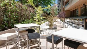 Une terrasse extérieure avec des tables et des chaises, entourée de verdure et bordée d’un bâtiment doté de grandes fenêtres.