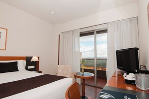 Ein Hotelzimmer mit Bett, Schreibtisch mit Fernseher, kleinem Tisch mit Pflanze und Blick auf die Stadt durch das Fenster.