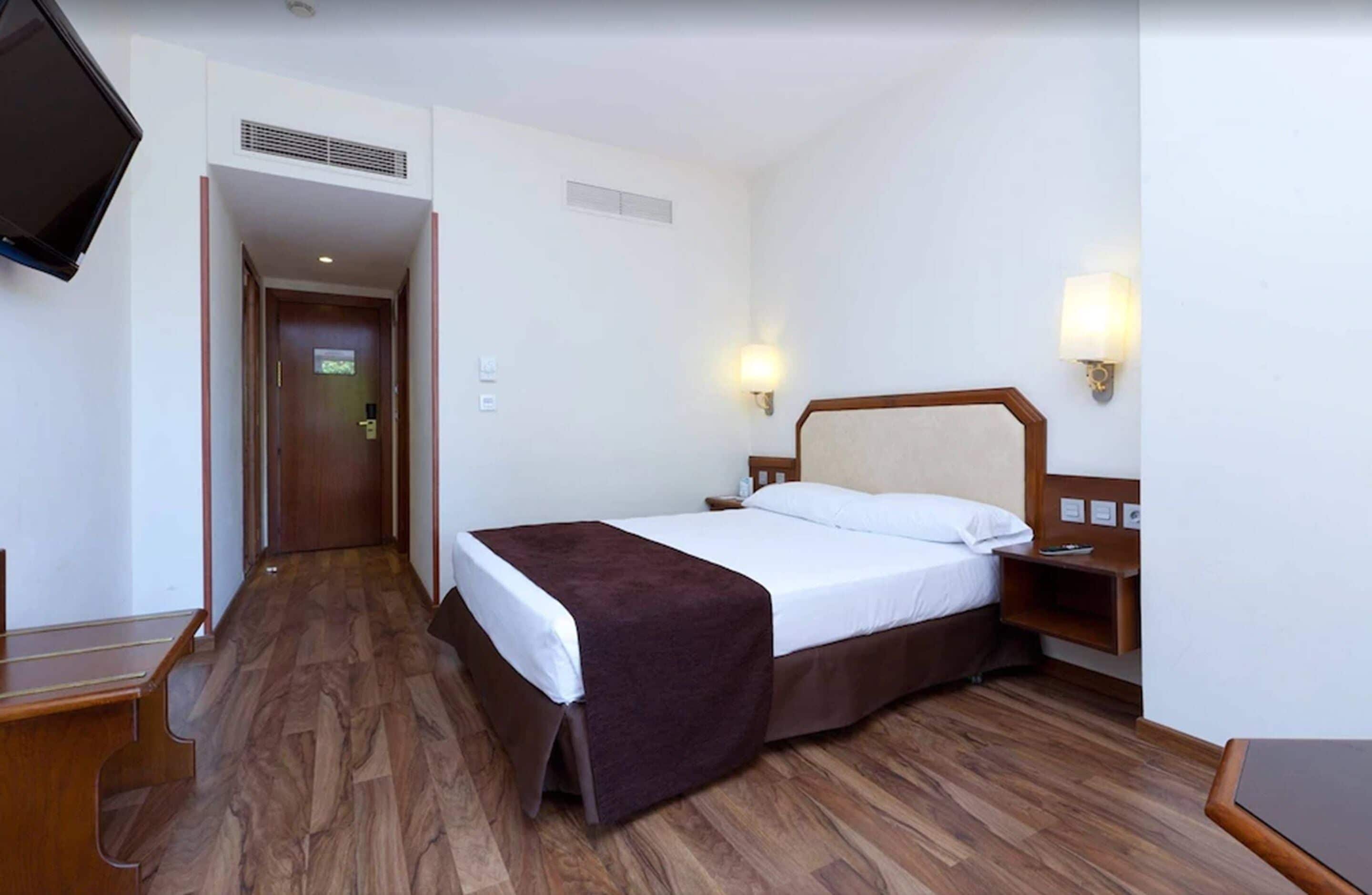 Habitación de hotel con piso de madera, cama con cabecera marrón, televisor montado en la pared y dos lámparas de pared.