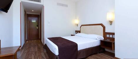 Habitación de hotel con piso de madera, cama con cabecera marrón, televisor montado en la pared y dos lámparas de pared.