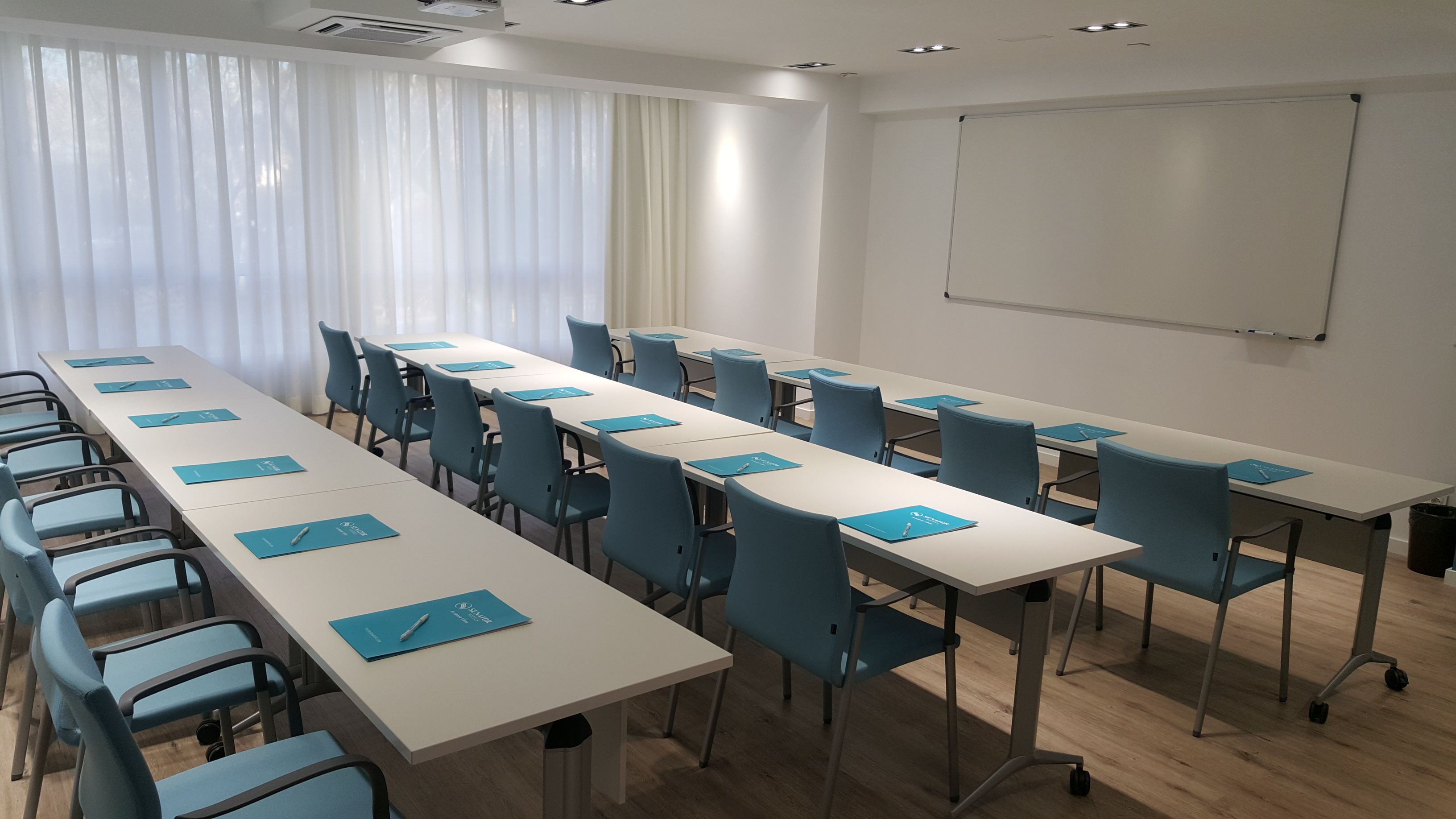 Une salle de conférence avec de longues tables, des chaises bleues et un tableau blanc au mur.