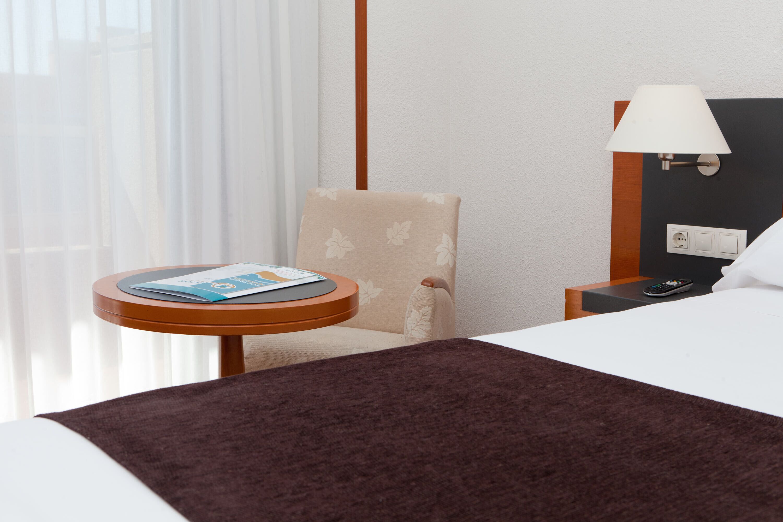 Una habitación de hotel con una cama, una mesita con una revista, una silla, una lámpara y una mesita de noche con un control remoto.