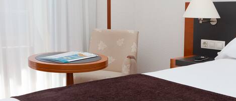 Una habitación de hotel con una cama, una mesita con una revista, una silla, una lámpara y una mesita de noche con un control remoto.