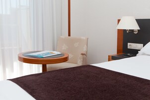 Ein Hotelzimmer mit einem Bett, einem kleinen Tisch mit einer Zeitschrift, einem Sessel, einer Lampe und einem Nachttisch mit Fernbedienung.