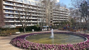 Ein öffentlicher Park mit einem runden Springbrunnen, umgeben von violetten Blumen, Bänken und Spielgeräten.