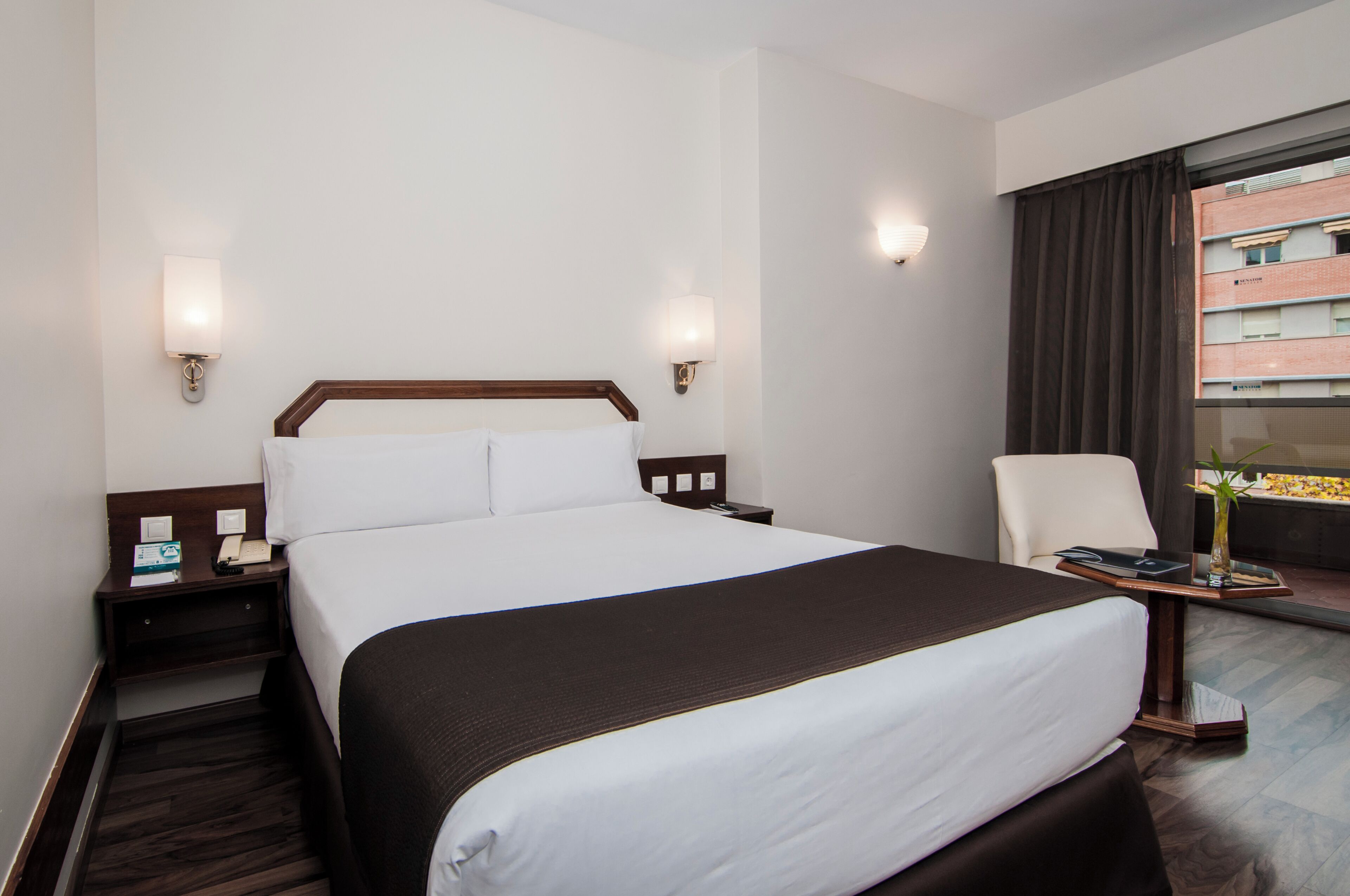 Habitación de hotel con una cama grande, mesitas de noche, una silla, una pequeña mesa con un florero y una ventana con cortinas.