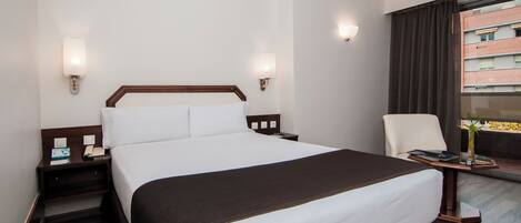 Habitación de hotel con una cama grande, mesitas de noche, una silla, una pequeña mesa con un florero y una ventana con cortinas.