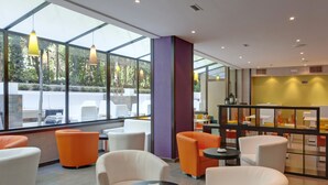 Un espace de détente intérieur moderne, avec des chaises blanches et orange, un mur en verre donnant sur un espace verdoyant et un mur d’accent violet.
