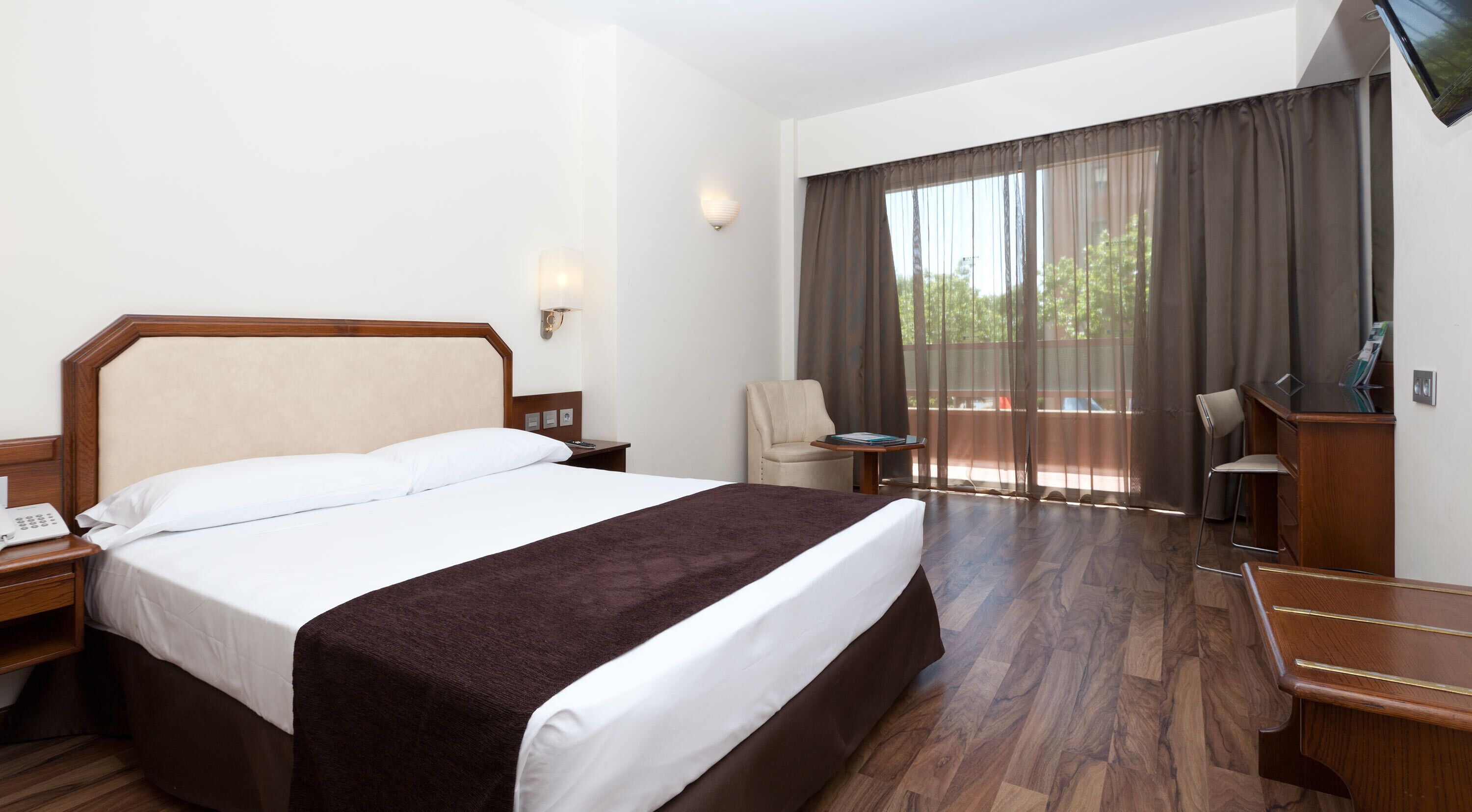 Habitación de hotel con una cama grande, un escritorio, una silla y vistas al exterior a través de una ventana con cortinas.