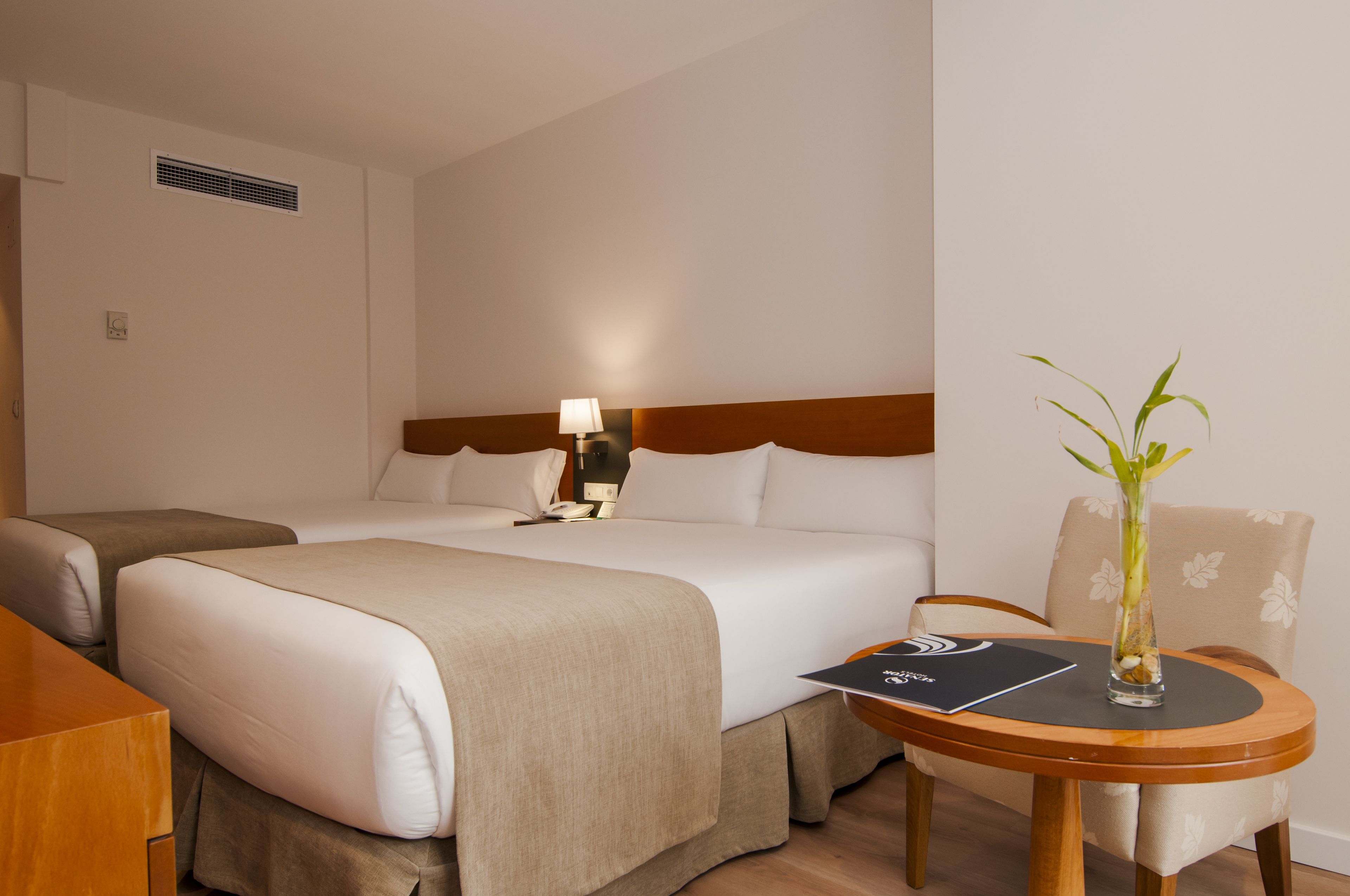 Une chambre d’hôtel avec deux lits, une table de chevet avec une lampe, une chaise et une petite table sur laquelle est posé un vase.