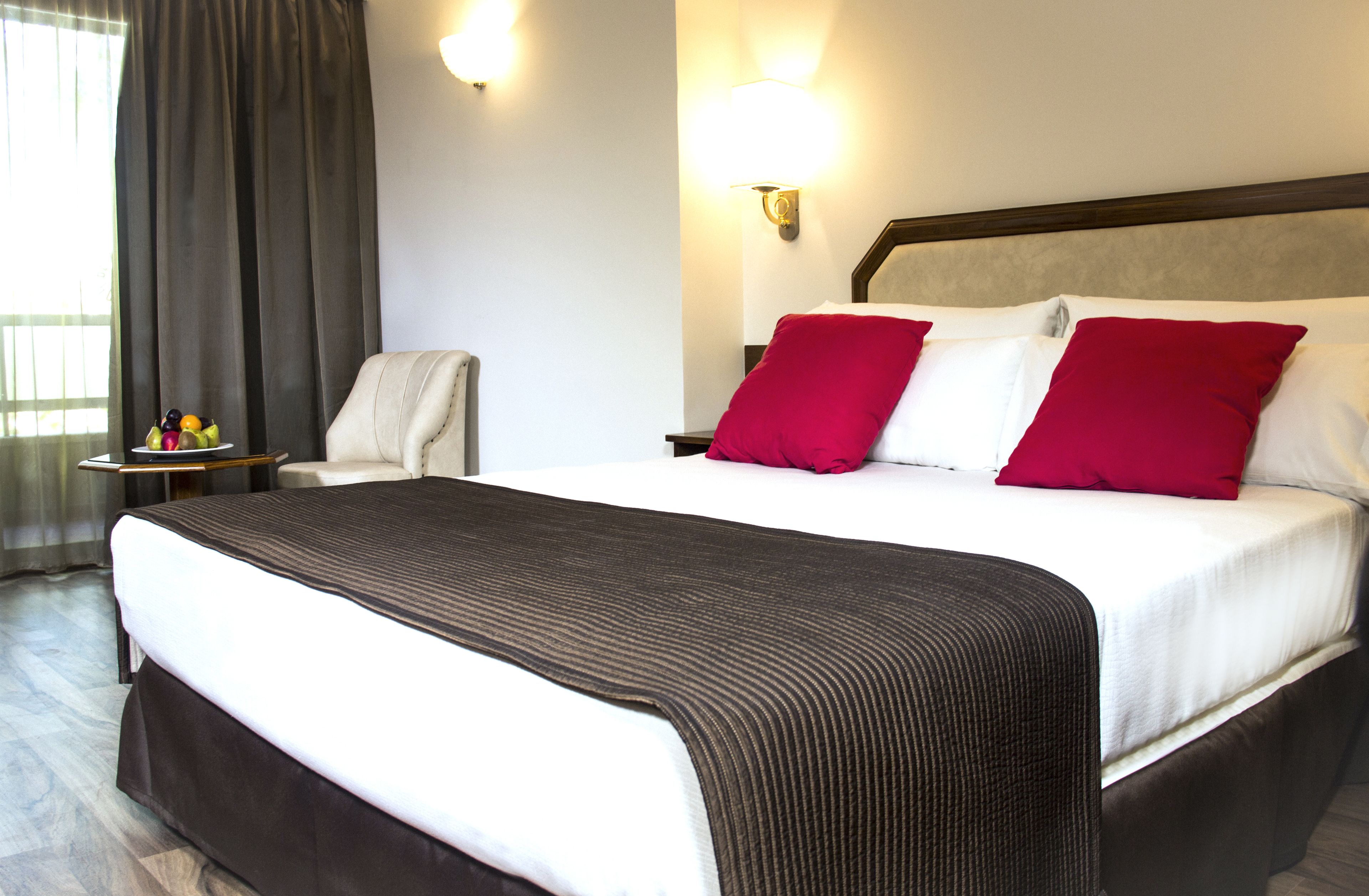 Une chambre d’hôtel avec un grand lit, deux oreillers rouges, une couette blanche, une couvre-lit marron, une table de chevet avec une lampe et une chaise.